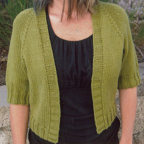 A person is wearing the Summer Open Cardigan by Bryson Distributing, Inc., a knitted green short-sleeve summer cardigan, over a black top. Made with worsted weight yarn, the Summer Open Cardigan features ribbed edges along the front opening, sleeves, and hem for an easy-fitting look. The person's head is not fully visible in the image. The background features greenery and a stone wall.