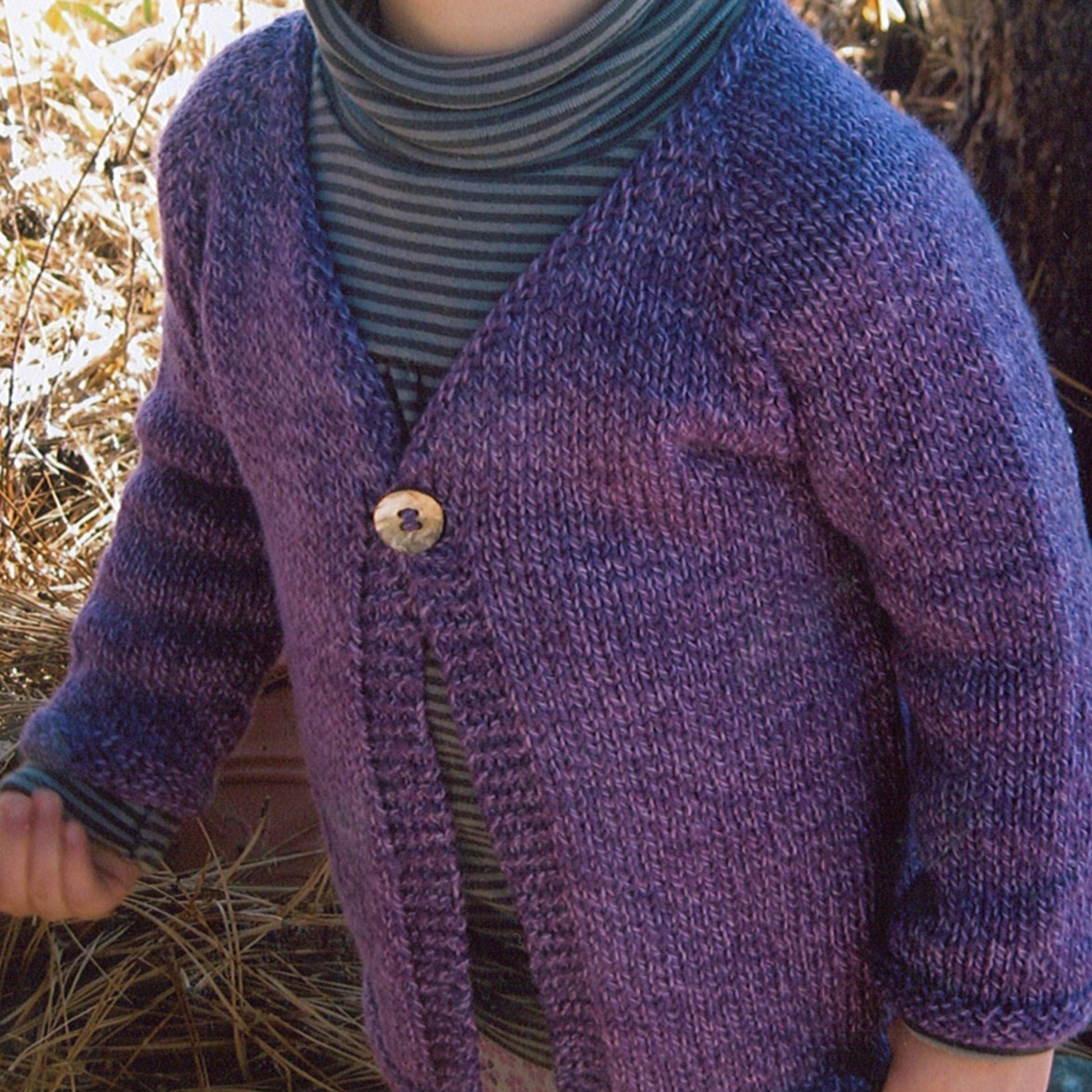 A young child wearing Bryson Distributing, Inc.'s Girl's One Button Cardigan in purple and a striped turtleneck stands outdoors in a wooded area, with sunlight filtering through the trees. Surrounded by dried leaves and branches, the scene beautifully captures the charm of children enjoying nature.