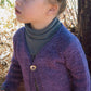 A young child with curly hair, tied back, is standing outdoors, gazing upwards. They are wearing a Bryson Distributing, Inc.'s Girl's One Button Cardigan in purple over a gray striped turtleneck. The background features dried grass, branches, and a tree trunk. The child's lightweight sweater seems cozy as they wear a thoughtful expression.