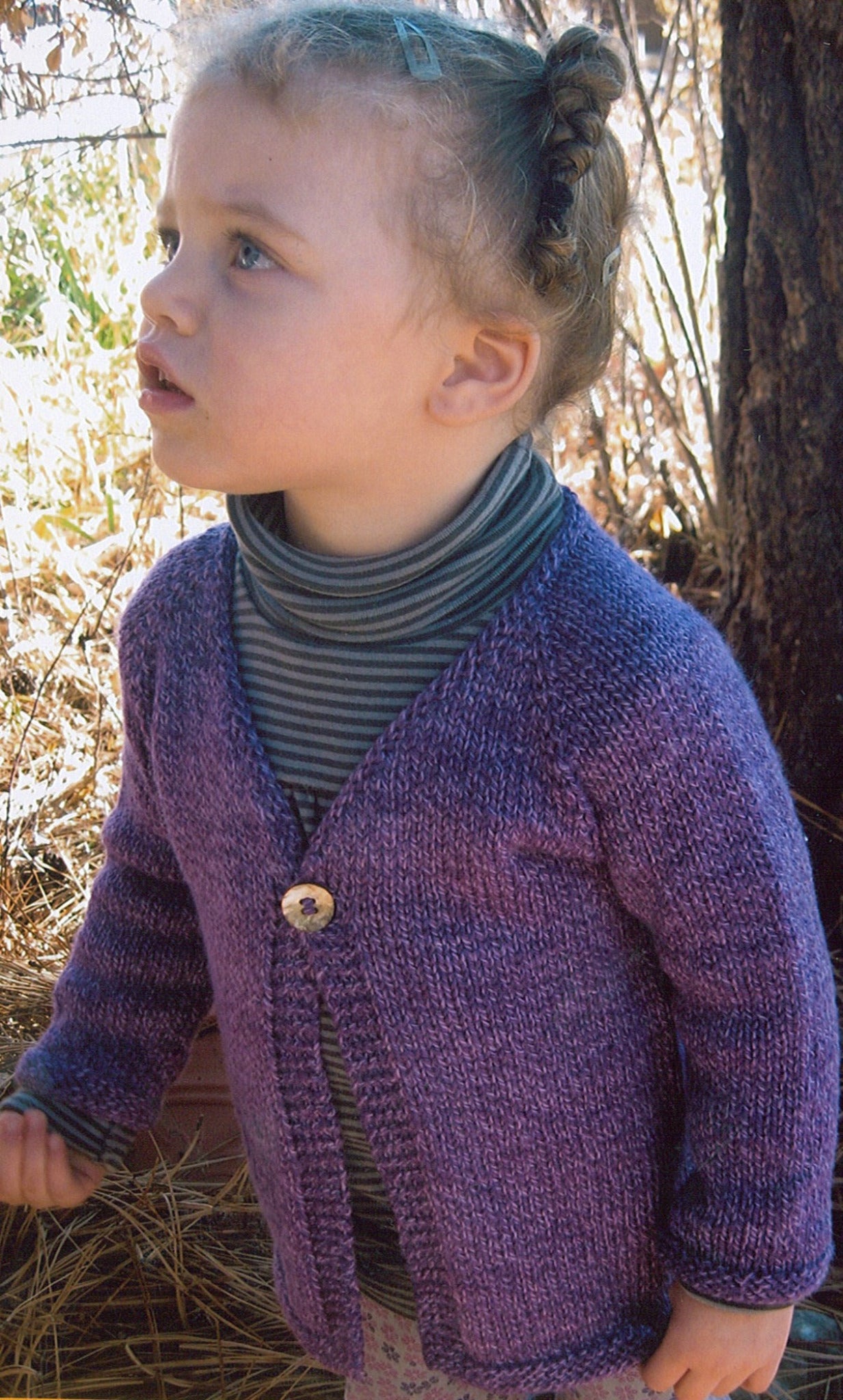 A young child with curly hair, tied back, is standing outdoors, gazing upwards. They are wearing a Bryson Distributing, Inc.'s Girl's One Button Cardigan in purple over a gray striped turtleneck. The background features dried grass, branches, and a tree trunk. The child's lightweight sweater seems cozy as they wear a thoughtful expression.