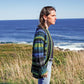 A person with shoulder-length hair stands in profile, gazing at the ocean. They wear a Top Down Shawl Collar Cardigan by Bryson Distributing, Inc., made of variegated yarns, paired with jeans. The background features a sunny coastal landscape with grassy areas and a clear blue sky over the sea.