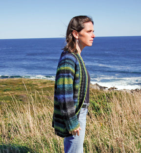 A person with shoulder-length hair stands in profile, gazing at the ocean. They wear a Top Down Shawl Collar Cardigan by Bryson Distributing, Inc., made of variegated yarns, paired with jeans. The background features a sunny coastal landscape with grassy areas and a clear blue sky over the sea.