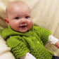 A smiling baby, fair-skinned and round-faced, sits against a soft cream-colored blanket, wearing the vibrant green Lacy Baby Hoodie by Bryson Distributing, Inc. The hoodie features a three-button closure and a delicate lace edge. The baby gazes directly into the camera with curiosity and charm.