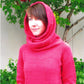 A person stands facing forward, wearing the Top Down Cowl Hoodie by Knitting from Bryson Distributing, Inc., which has a red color and a furry hood trim. The background features a green-leafed tree and a white wall. The person appears to be outdoors and is gazing slightly to the right.