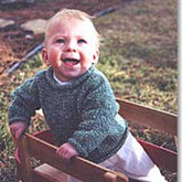 A smiling baby with light-colored hair is wearing a green sweater called the "Baby Neck Down Pullover" by Bryson Distributing, Inc. and white pants. The baby is outdoors, leaning over the side of a small wooden wagon with a grassy and slightly blurred background, creating a bright and cheerful scene.