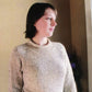 A person with short dark hair stands by a window, looking thoughtfully into the distance. They are wearing a beige knitted New Neckdown Pullover for Women from Bryson Distributing, Inc. The background is softly lit, featuring a yellow wall and a shadowy glimpse of a plant or flowers.