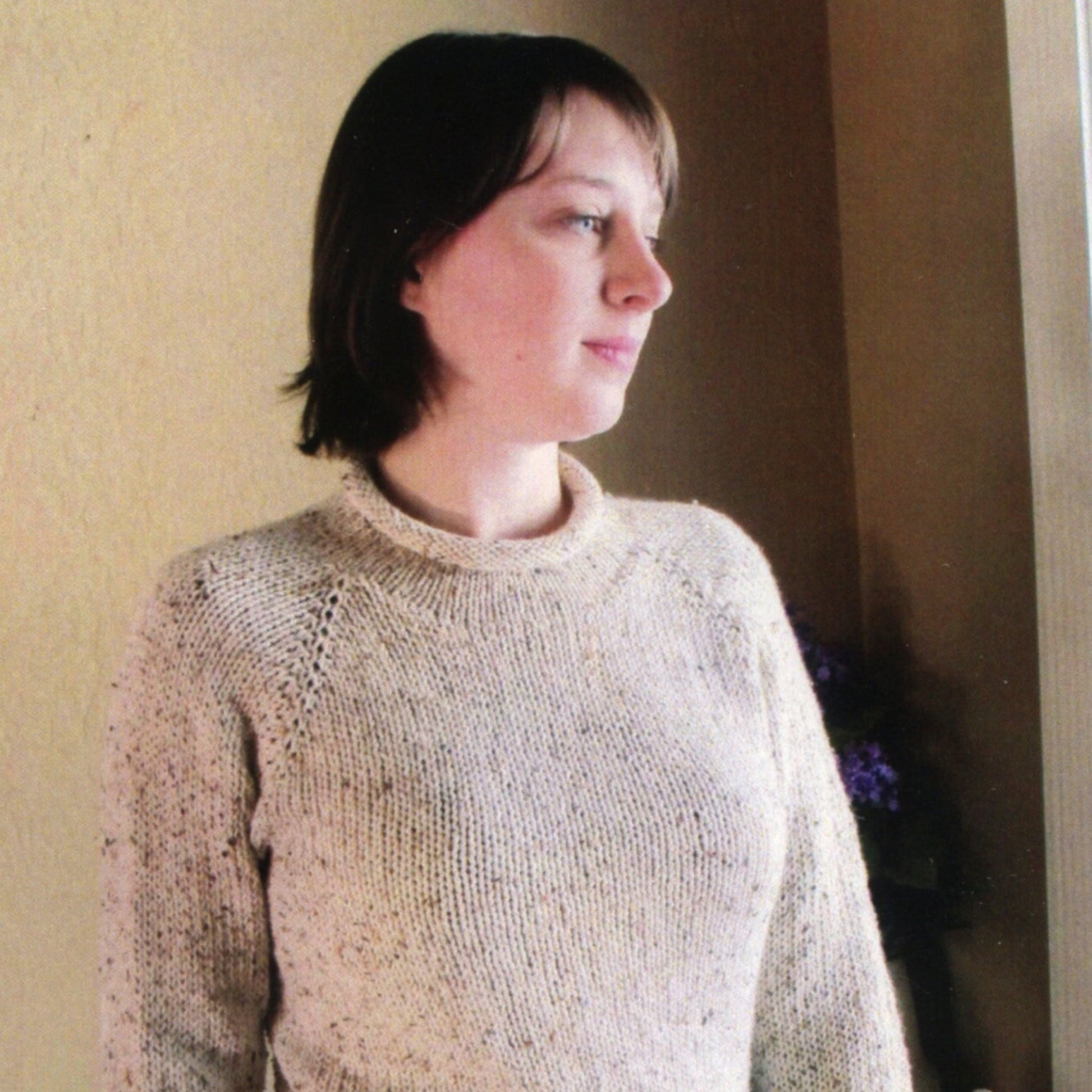 A person with short dark hair stands by a window, looking thoughtfully into the distance. They are wearing a beige knitted New Neckdown Pullover for Women from Bryson Distributing, Inc. The background is softly lit, featuring a yellow wall and a shadowy glimpse of a plant or flowers.