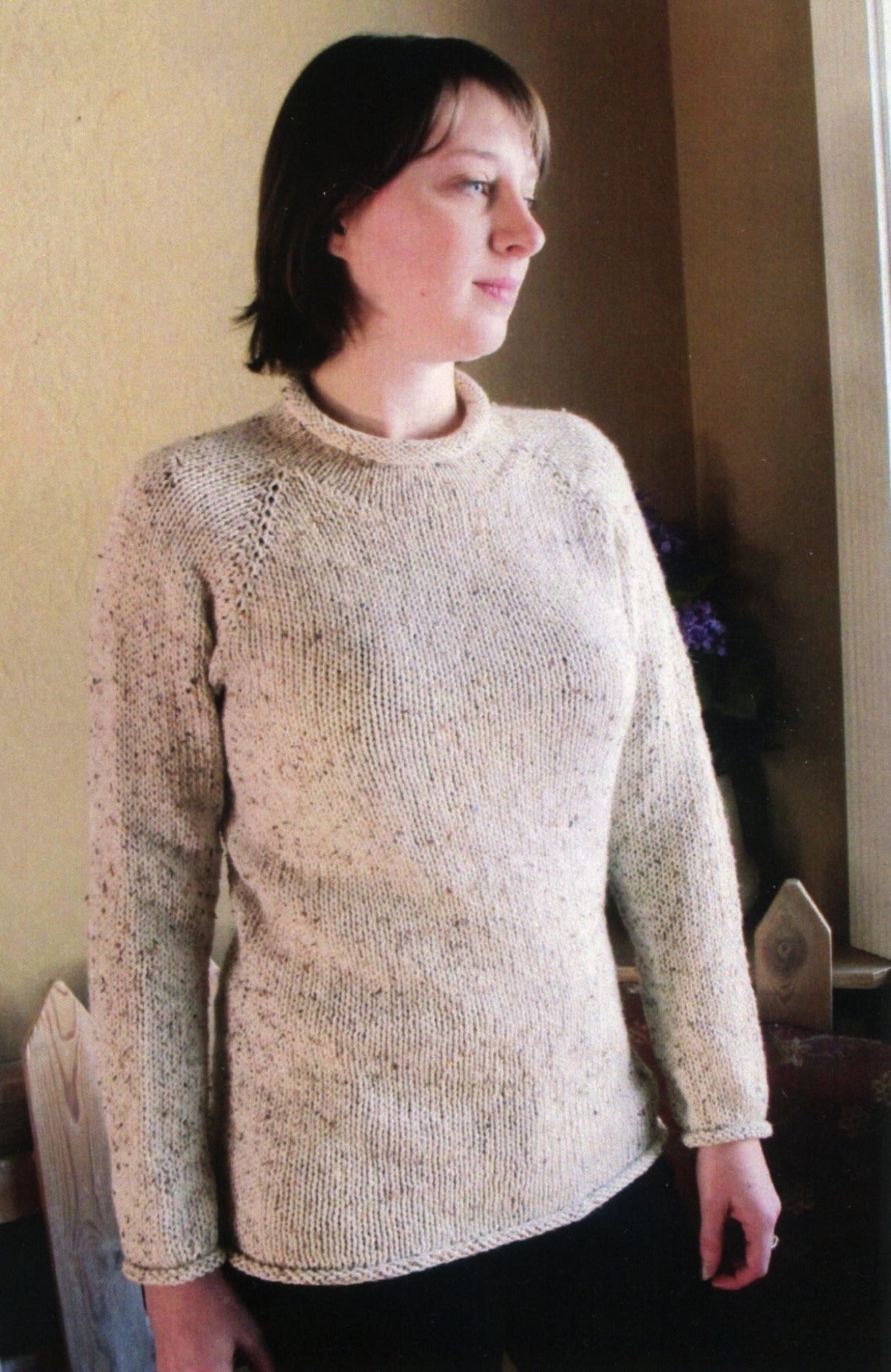 A person with short brown hair stands indoors wearing a light-colored, long-sleeve New Neckdown Pullover for Women, designed by Bryson Distributing, Inc. for a flattering fit. They are facing slightly to the right and looking into the distance. The background features a beige wall and some indistinct objects.