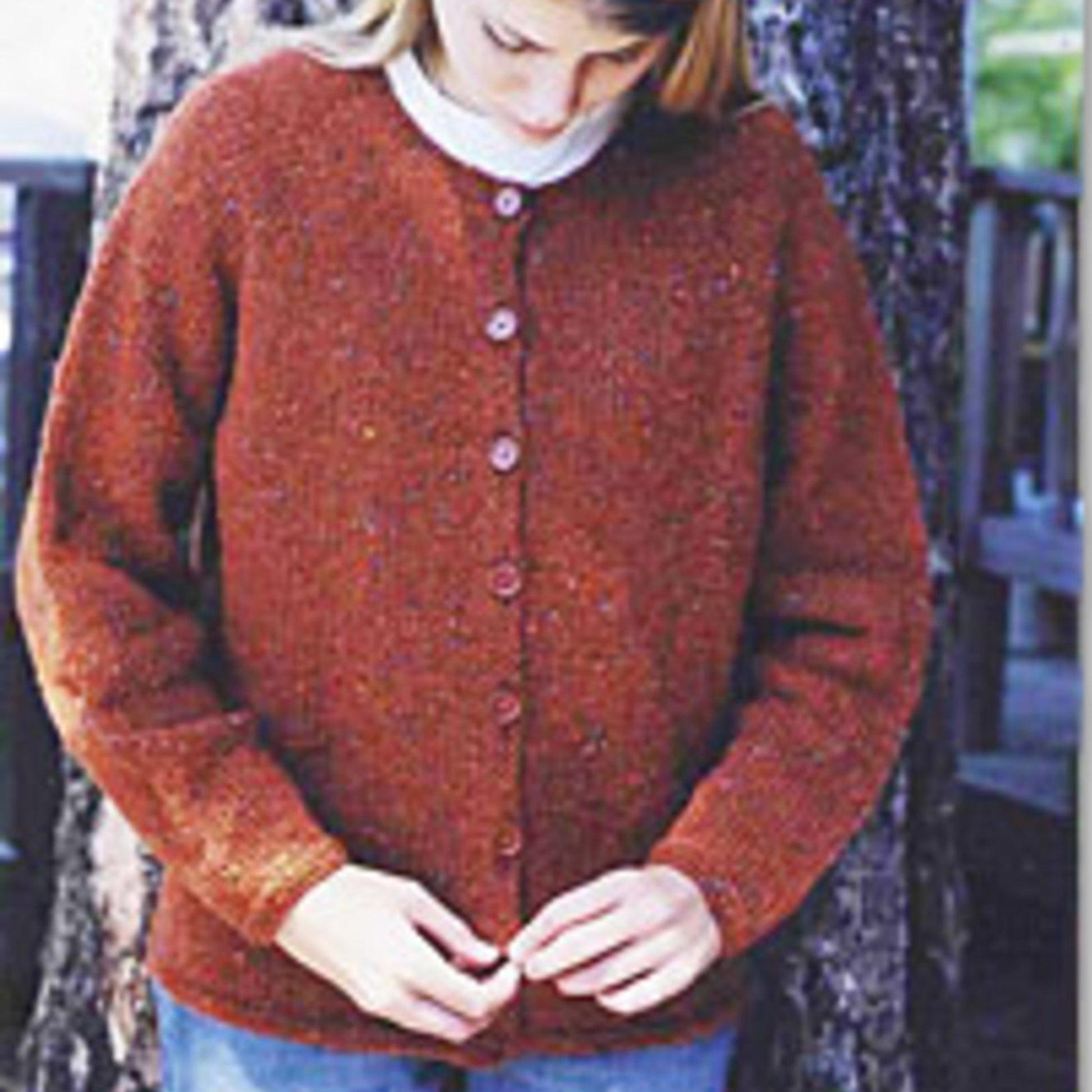 A person with light brown hair, head slightly tilted downwards, is buttoning a maroon Bryson Distributing, Inc. Neck Down Cardigan featuring garter stitch borders. They are standing outdoors in front of a tree and are wearing blue jeans and a white shirt beneath the cardigan.