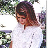 A woman with long blonde hair, wearing a white Henley T-shirt from Bryson Distributing, Inc., is looking down with a slightly serious expression. She stands outdoors in front of some greenery and a wooden fence, the comfy Bryson T-shirt perfectly complementing the serene, natural setting.