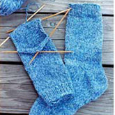 Bryson Distributing, Inc.'s Beginner Socks in a lovely blue color are in progress on double-pointed needles, displayed on a wooden surface. One sock is fully complete, while the other is partially finished with yarn and needles still attached. A ball of blue yarn can also be seen.