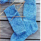 The product "Beginner Socks" by Bryson Distributing, Inc. features two blue women's socks on a wooden surface. One sock is fully finished while the other remains partially knitted, with a ball of matching variegated blue yarn and several wooden knitting needles still attached, adding an artisanal touch to these otherwise basic socks.