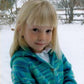 A young girl with light blonde hair and straight bangs is wearing a Bryson Distributing, Inc. Children's Neck Down Cardigan in a green and blue pattern. She is outdoors in a snowy area, surrounded by trees and a wooden fence in the background. The girl is looking at the camera with a slight smile.