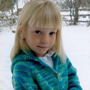 A young girl with light blonde hair and straight bangs is wearing a Bryson Distributing, Inc. Children's Neck Down Cardigan in a green and blue pattern. She is outdoors in a snowy area, surrounded by trees and a wooden fence in the background. The girl is looking at the camera with a slight smile.