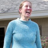 A woman with light hair tied back is standing outside in front of a building with gray roof shingles and beige siding. She is wearing a light blue Bulky Neck Down Pullover from Bryson Distributing, Inc., made of chunky yarn, and is smiling broadly. The lighting suggests it is a sunny day.