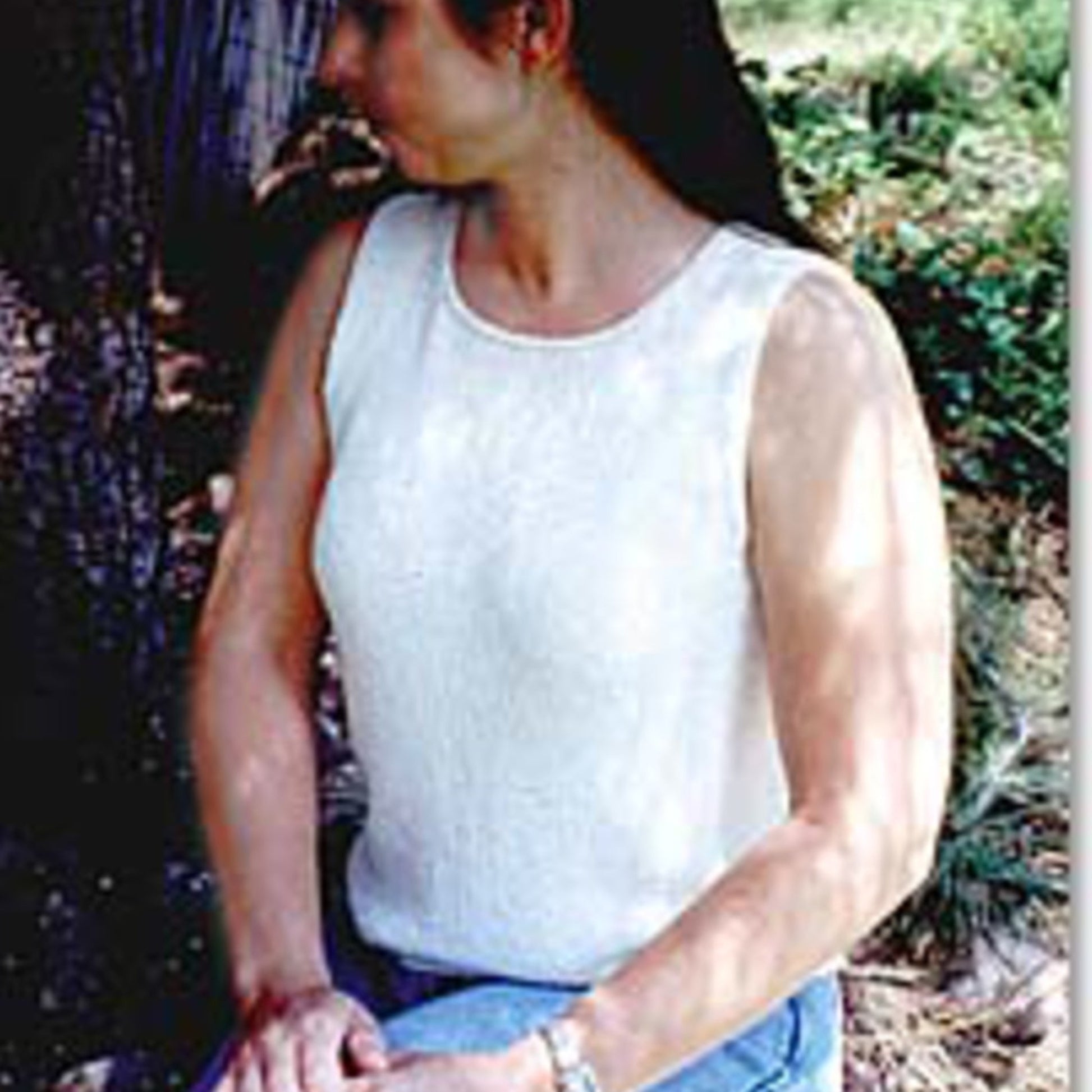 A woman wearing a sleeveless Tank Top from Bryson Distributing, Inc. and blue jeans is sitting outdoors on a bench. She is looking to her right, away from the camera, with her hands resting on her lap. Greenery and trees are visible in the background.