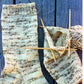 A close-up of hand-knitted Men's Heavy Weight Boot Socks by Bryson Distributing, Inc. on a wooden surface. On the left is a completed light-colored heavy boot sock with brown and beige accents, while on the right, another similar sock made from worsted weight yarn is still in progress with knitting needles and yarn attached. Suitable for men's shoe size 7 to 12.