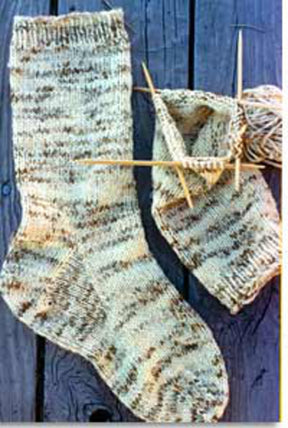 A completed Men's Heavy Weight Boot Sock from Bryson Distributing, Inc. with a beige and brown pattern rests on a blue wooden surface beside a second sock in progress. Crafted from worsted weight yarn, these heavy boot socks fit men's shoe sizes 7 to 12. Wooden knitting needles are inserted into the unfinished sock, while a ball of yarn lies adjacent.