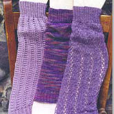 Display three leg warmers from the Bryson Distributing, Inc. Leg Warmers Pattern collection, each meticulously crafted using circular needles from soft yarn in various shades of purple. The one on the left features a light purple with a ribbed texture, the center pairs multiple hues of purple, and the right exhibits an intricate lacy pattern. Elegantly arranged on a wooden surface, they create a stunning visual display.