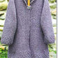 An "Autumn Coat" from Bryson Distributing, Inc. is displayed on a wooden hanger against a stone wall background. This gray, hand-knitted tunic-style cardigan with long sleeves features a simple, textured pattern and boasts a comfortable fit, fastened at the top with a decorative silver clasp.