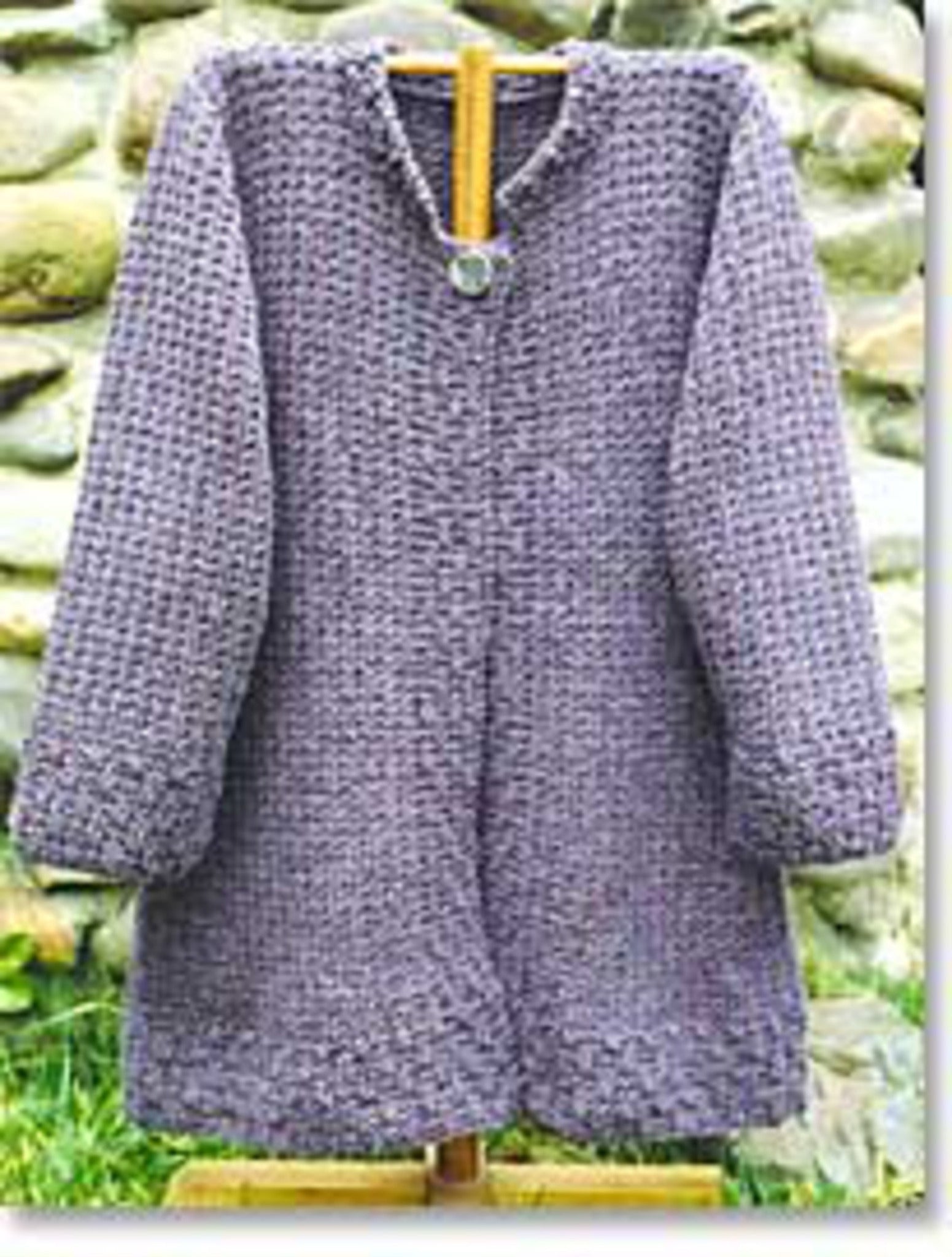 An "Autumn Coat" from Bryson Distributing, Inc. is displayed on a wooden hanger against a stone wall background. This gray, hand-knitted tunic-style cardigan with long sleeves features a simple, textured pattern and boasts a comfortable fit, fastened at the top with a decorative silver clasp.