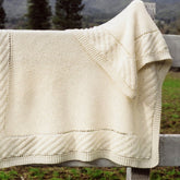 A cream-colored Prairie Blanket from Bryson Distributing, Inc., featuring a textured garter stitch pattern, is draped over a wooden fence. The background showcases blurred trees and a hint of a mountain under the gray sky, enhancing the cozy charm of this exquisite creation.