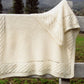 The Prairie Blanket from Bryson Distributing, Inc., a beige baby blanket with a textured pattern on the edges, crafted in a cozy garter stitch from soft DK yarn, is draped over a wooden fence in an outdoor setting. The background features blurred green grass and trees.