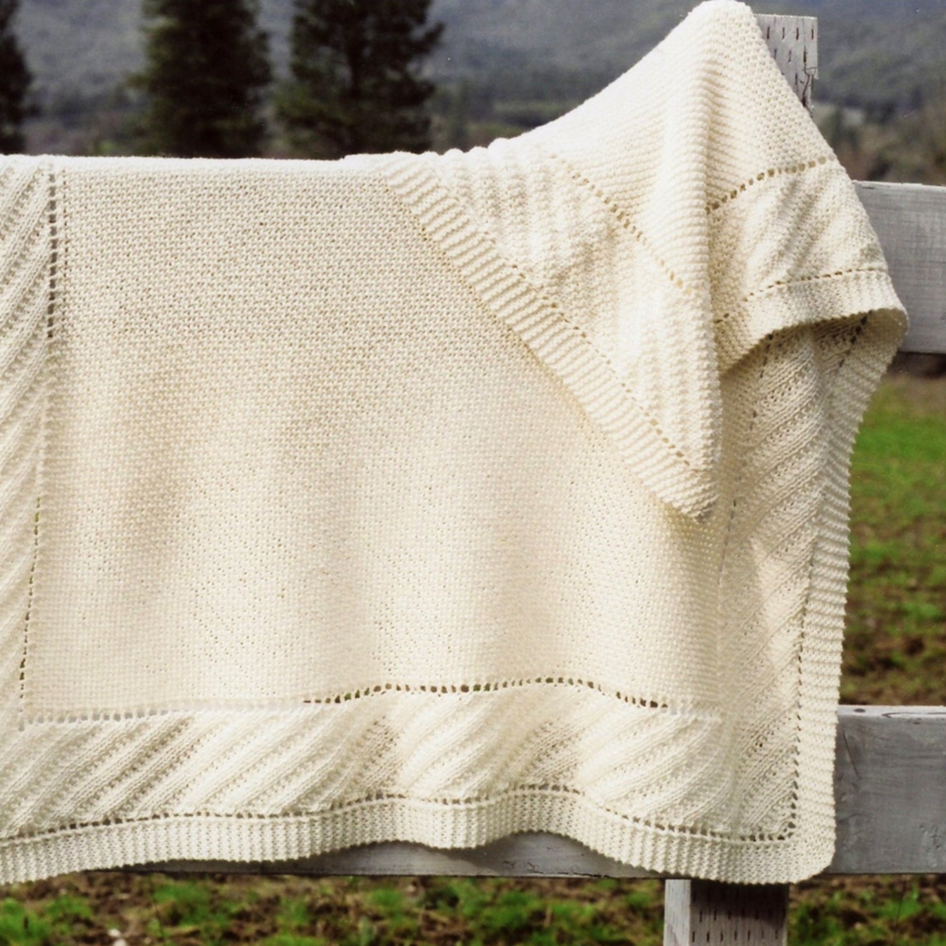 A cream-colored Prairie Blanket from Bryson Distributing, Inc., featuring a textured garter stitch pattern, is draped over a wooden fence. The background showcases blurred trees and a hint of a mountain under the gray sky, enhancing the cozy charm of this exquisite creation.