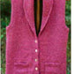 A knitted, pinkish-red cardigan with six buttons and two small front pockets from Bryson Distributing, Inc., named the Village Vest, hangs on a wooden hanger. The sleeveless design offers extra warmth while maintaining a tailored look. The background features a blurred natural setting with greenery.