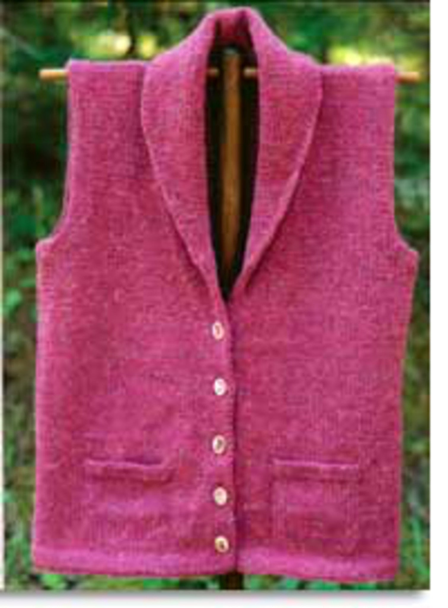 Displayed on a wooden hanger, the Village Vest Pattern by Bryson Distributing, Inc. exudes warmth with its knitted pink fabric and shawl collar. Featuring six buttons and two small pockets, this tailored piece stands out against the green outdoor background.