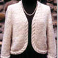 A beige, open-front Seville Cardigan/Jacket from Bryson Distributing, Inc., featuring a cable trim along the edges and sleeves, draped over a black top. Displayed on a mannequin against a stone wall background, this v-necked cardigan is complemented by a pearl necklace around the mannequin's neck.