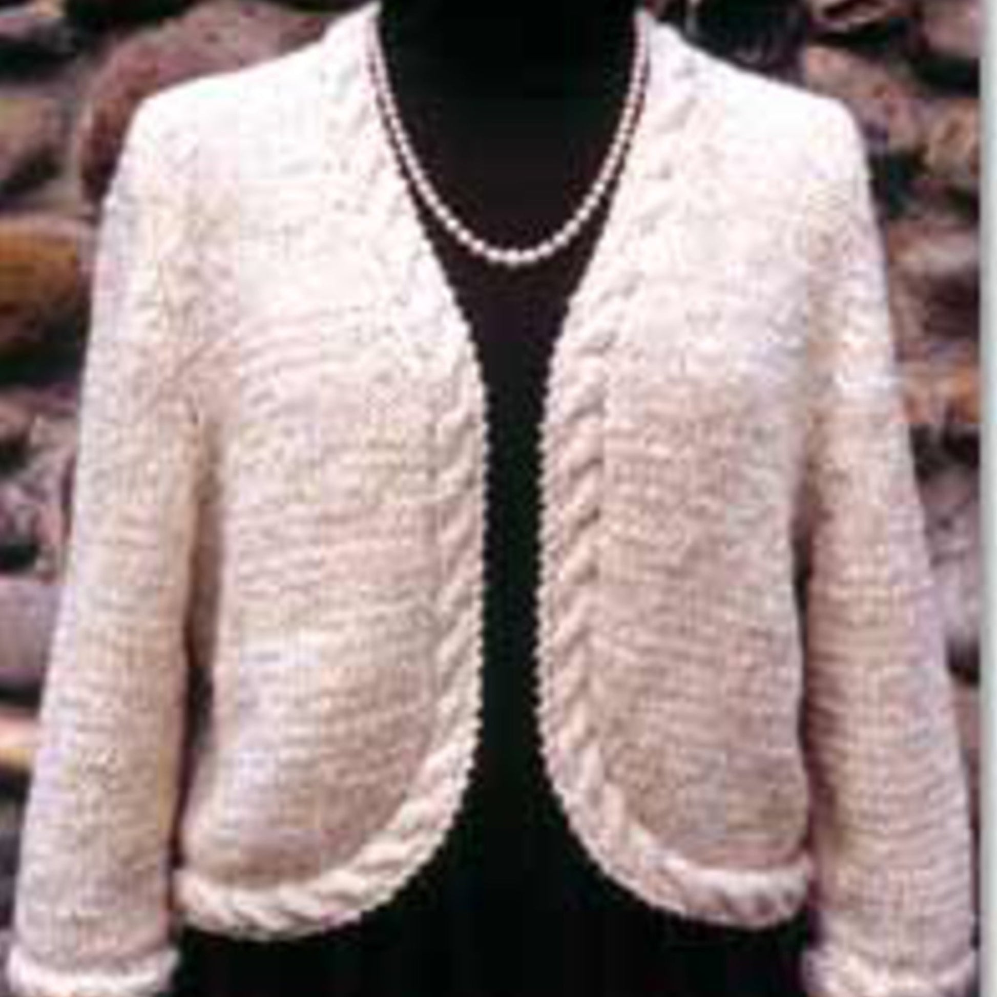 The Seville Cardigan/Jacket from Bryson Distributing, Inc. is showcased on a mannequin against a stone wall backdrop. This cream-colored garment displays an open-front style with braided trim along the edges and hem, exuding a simple yet elegant design featuring a round neckline.