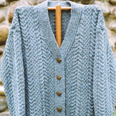 Hanging on a wooden hanger against a stone wall, the Caswell Cardigan by Bryson Distributing, Inc. features light blue knitted fabric with a textured zigzag pattern, wooden buttons, and a V-neck—a perfect favorite go-to sweater for cozy days.