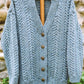 Hand-knitted Caswell Cardigan by Bryson Distributing, Inc. in gray Aran Cable design with a deep V-neckline, displayed on a wooden hanger against a stone wall background. The textured cardigan features five intricately designed buttons down the front.