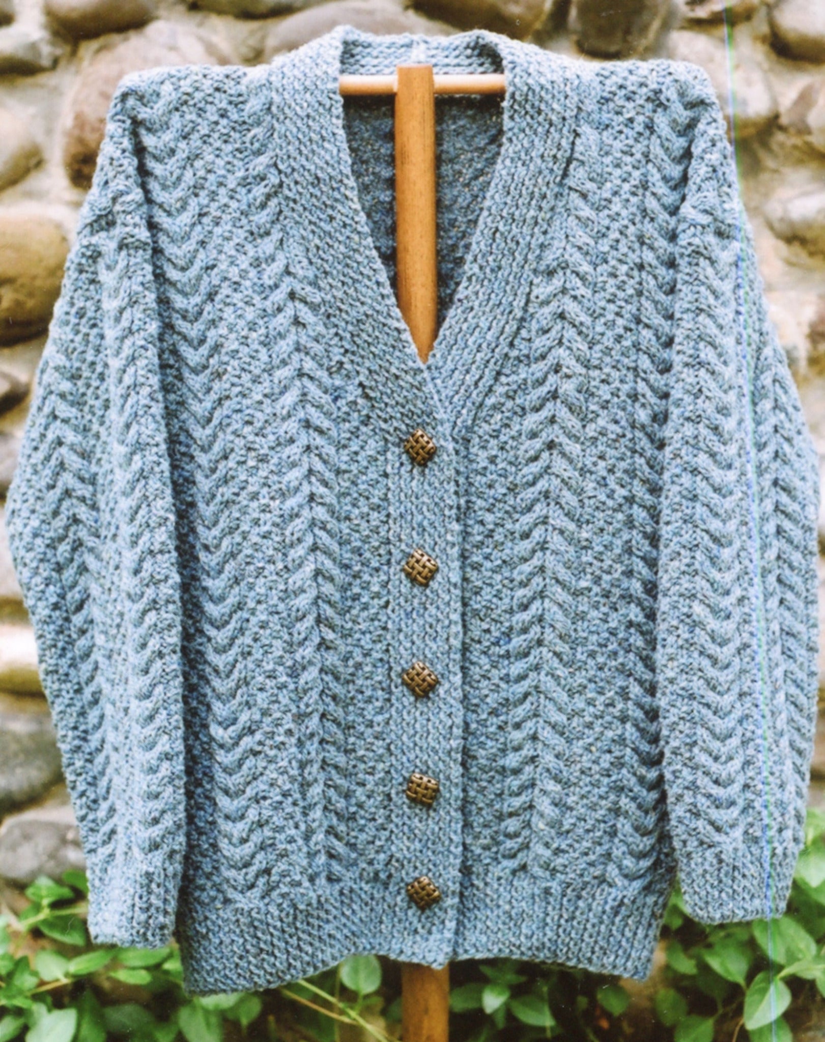 Hand-knitted Caswell Cardigan by Bryson Distributing, Inc. in gray Aran Cable design with a deep V-neckline, displayed on a wooden hanger against a stone wall background. The textured cardigan features five intricately designed buttons down the front.