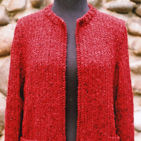 A close-up of a red Montreal Jacket by Bryson Distributing, Inc. displayed on a dark mannequin. The jacket, open at the front with a round neckline and visible texture, could easily transition from an office setting to a night out. The background is out-of-focus and made up of large, rounded stones.