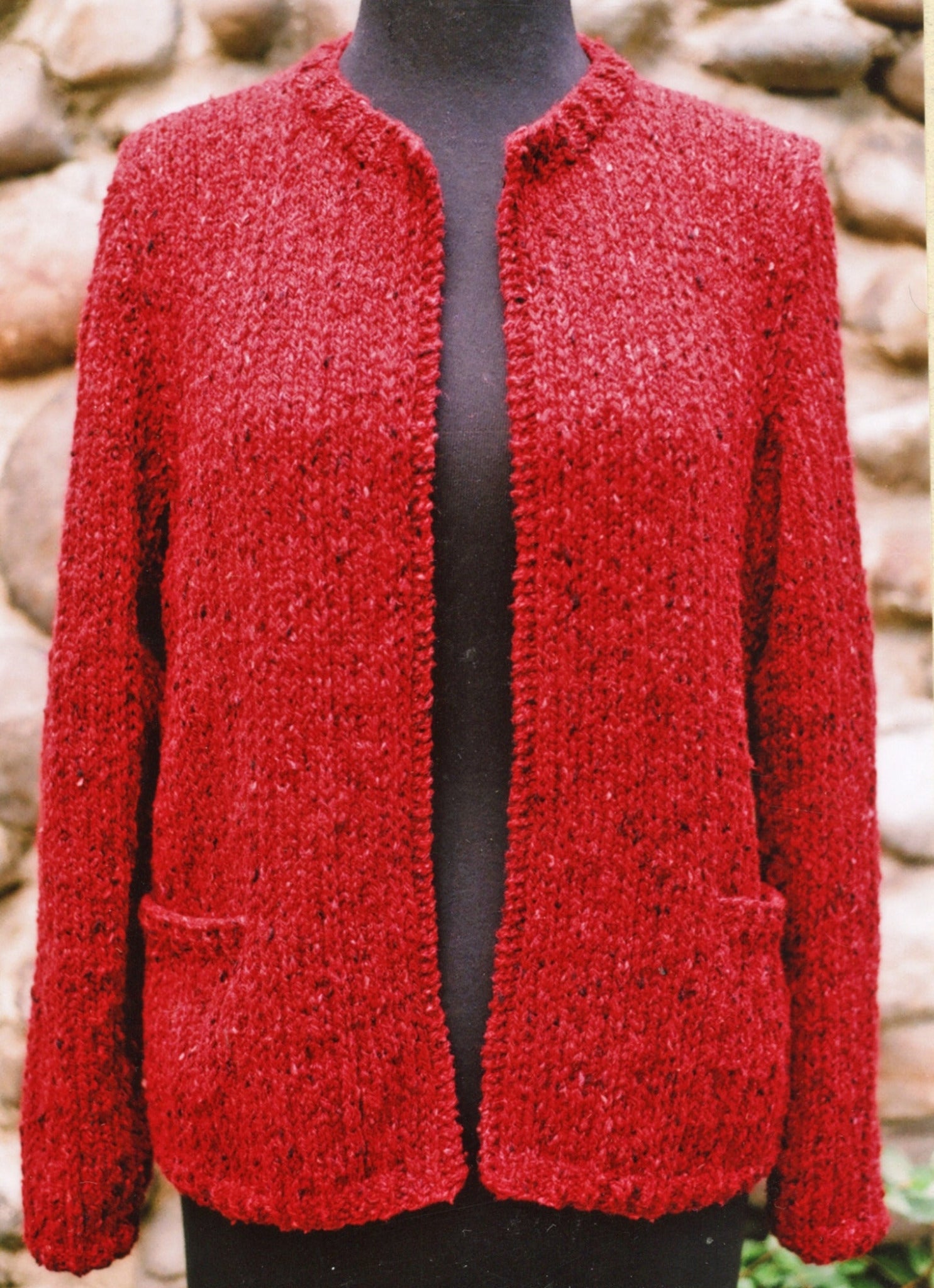 The Montreal Jacket by Bryson Distributing, Inc. is showcased on a black mannequin, ideal for both a night out and office wear. This red knitted cardigan features a straightforward, open front design without buttons and includes two front pockets. The background consists of a stone wall.