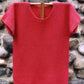 The Barbados Tee Pattern by Bryson Distributing, Inc. is displayed on a wooden hanger against a stone wall backdrop, showcasing its short-sleeved, red crocheted design made with delicate fingering yarn.