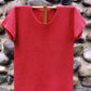 The Barbados Tee Pattern by Bryson Distributing, Inc. is shown on a wooden hanger, featuring its textured design and short sleeves in striking red made from fingering yarn. A rustic stone wall serves as the backdrop, accentuating the garment's vibrant color and intricate knitted pattern.