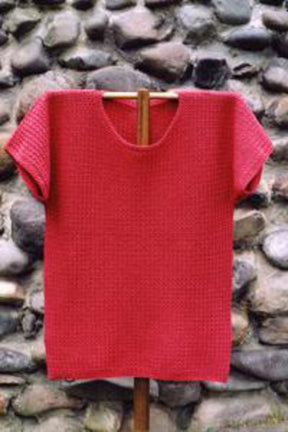 The Barbados Tee Pattern by Bryson Distributing, Inc. is shown on a wooden hanger, featuring its textured design and short sleeves in striking red made from fingering yarn. A rustic stone wall serves as the backdrop, accentuating the garment's vibrant color and intricate knitted pattern.