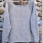 The West Side Raglan, a handmade, long-sleeved gray sweater from Bryson Distributing, Inc., is displayed against a stone wall backdrop. The tunic-style garment features a square neckline and is draped over a wooden hanger, highlighting its knitted texture and minimalist design.