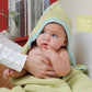 A baby wrapped in a vibrant Hooded Baby Blanket from Blue Sky Fibers, crafted with worsted weight cotton, is being cradled by an adult's hand. The baby has the blanket partially over its head and is touching its mouth with one finger. Behind them, there's a window and a red wall, while the Blue Sky Fibers logo is visible in the corner.