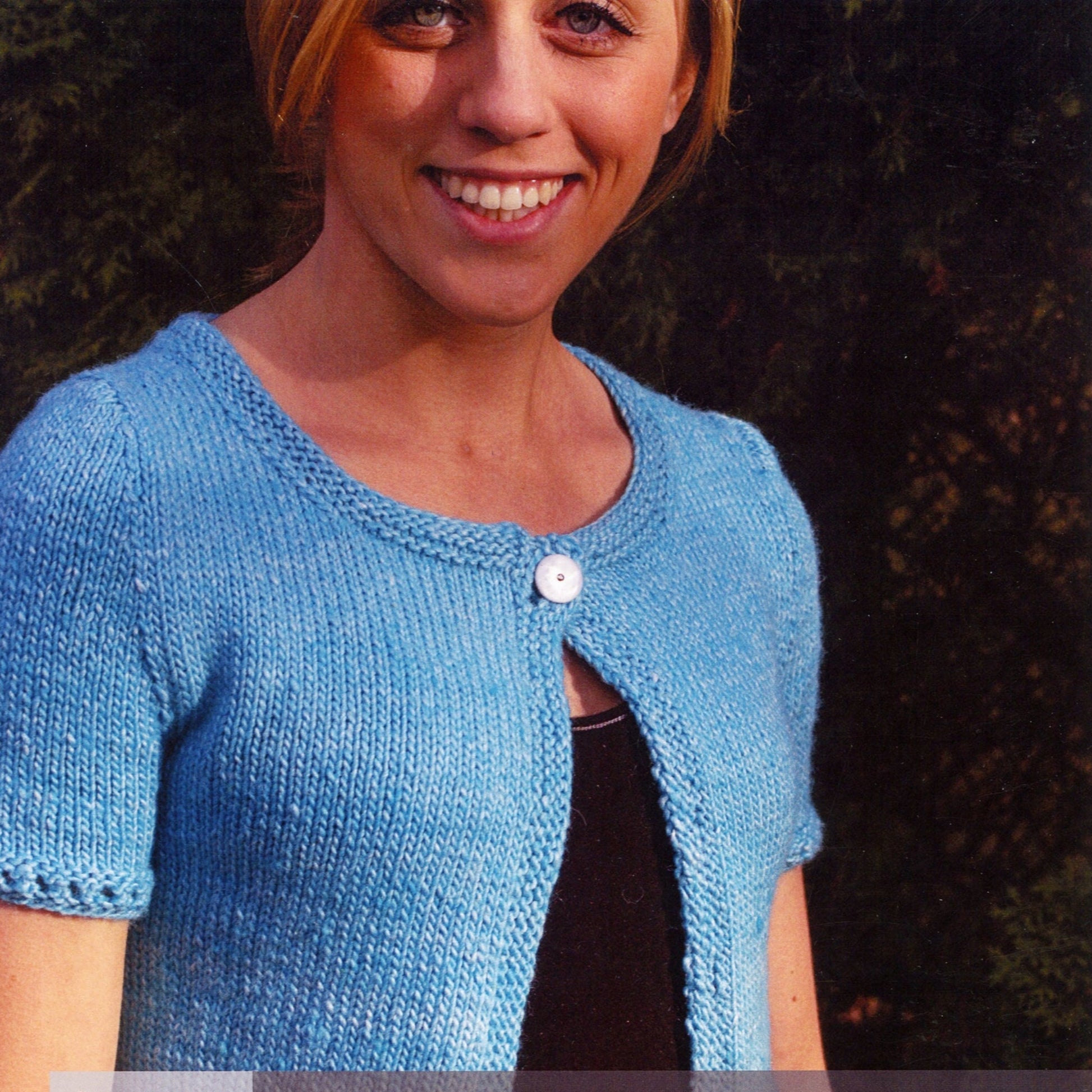 A person with short light hair, smiling, is wearing the Minty Short-Sleeved Cropped Cardi in light blue from Knitting Fever / Euro Yarns over a dark top. The background features dark green foliage.