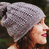 A person with curly hair and red lipstick is shown in profile view, wearing the chunky-knit Hedgerow Hat from The Crochet Collection by Knitting Fever / Euro Yarns, exuding vintage charm. The background is blurred, highlighting the detailed texture of the hat. The person is outdoors, suggesting a cool or wintery setting.