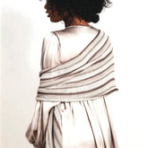 A person with curly hair facing away from the camera is wearing a light, flowing dress and a luxurious piece—a Blue Sky Fibers' Windom Silk Wrap draped diagonally over their shoulder. The wrap is striped in neutral tones, adding an air of everyday glamour against the plain white background.