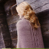 A person with long, wavy blond hair stands facing a weathered wooden wall. They are wearing the Hermite Poncho by Knitting Fever/Euro Yarns, featuring a thick, knitted gray design with a cable-knit pattern running down the back and a ribbed turtleneck collar.