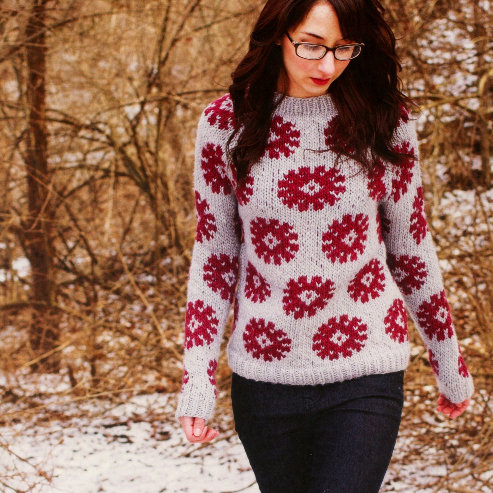 A woman strolls outside in the Supermoon Pullover Pattern by Knitting Fever / Euro Yarns, paired with dark jeans and glasses. The snowy, wooded backdrop enhances her cozy look.