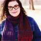 A person with long curly hair and glasses smiles while wearing a denim jacket and a colorful Squish Brioche Scarf from Knitting Fever / Euro Yarns. The background is blurred, suggesting an outdoor setting in daylight.