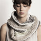 A person with closed eyes, straight bangs, and a serene expression is wearing the Caledonia Cowl from Blue Sky Fibers—a large, chunky knit scarf in light, neutral tones with subtle dark stripes. Made from organic cotton, this versatile cowl beautifully complements their sleeveless top against a plain, light-colored background.