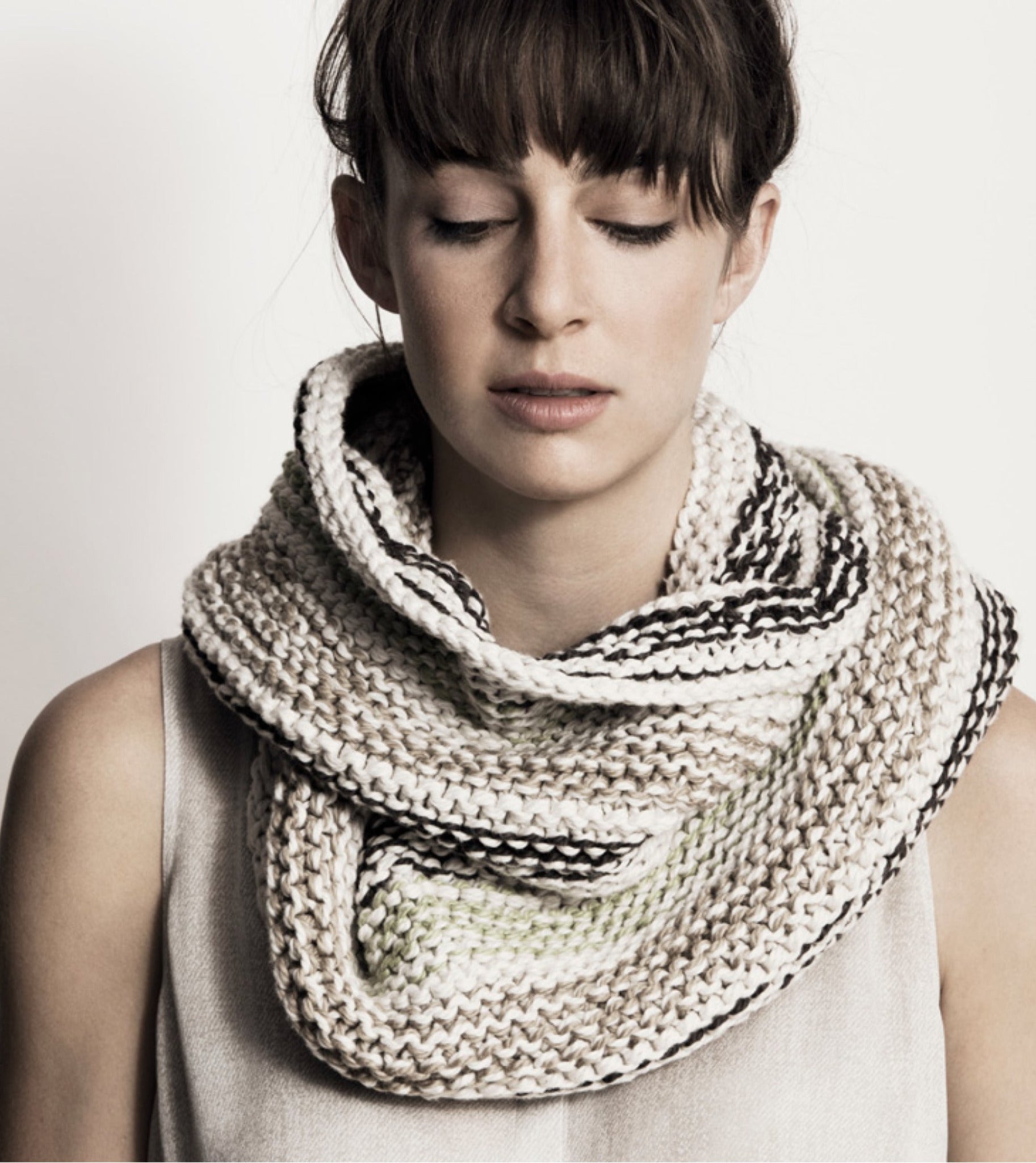 A person with closed eyes, straight bangs, and a serene expression is wearing the Caledonia Cowl from Blue Sky Fibers—a large, chunky knit scarf in light, neutral tones with subtle dark stripes. Made from organic cotton, this versatile cowl beautifully complements their sleeveless top against a plain, light-colored background.